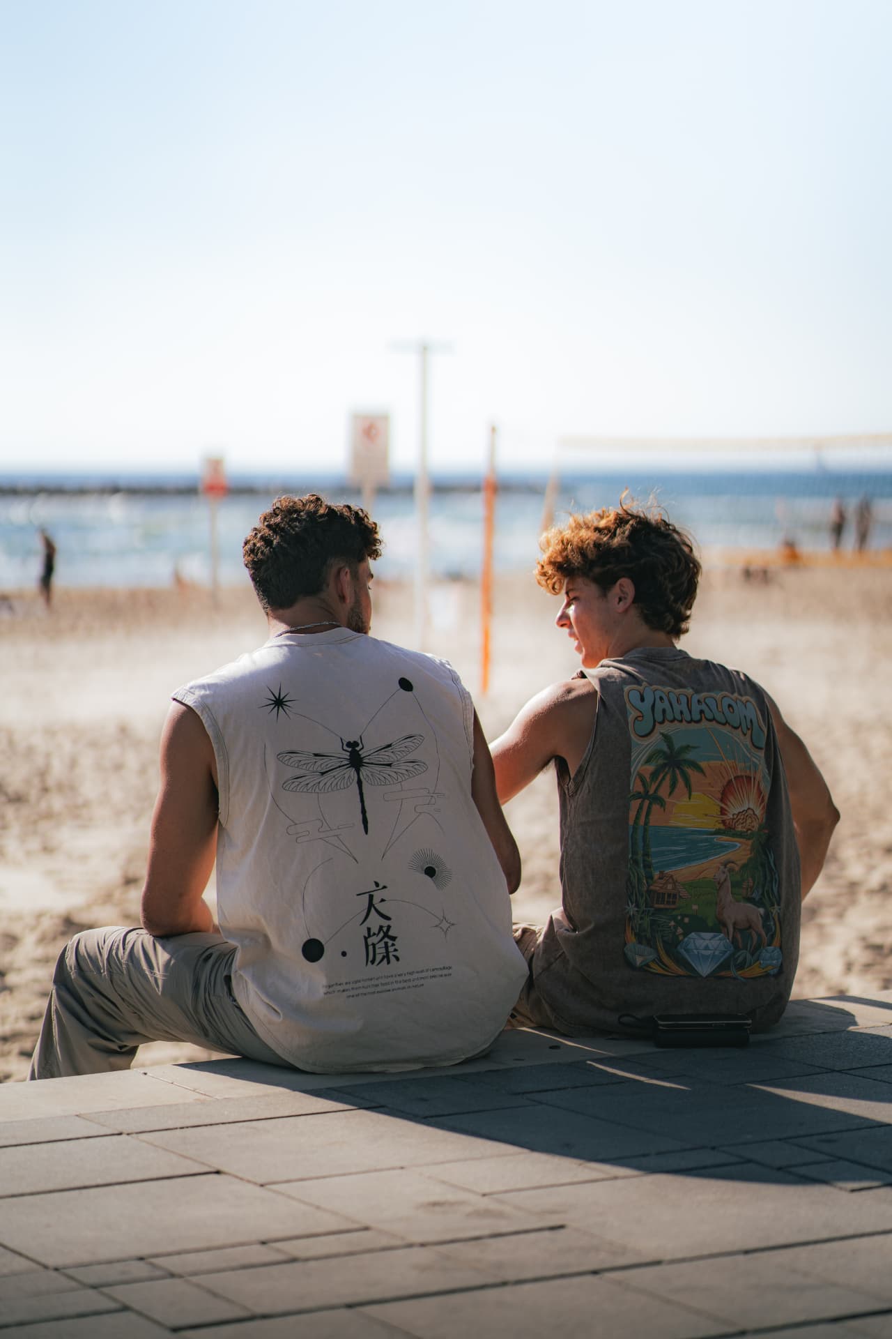 שני חברים יושבים על הטיילת בחוף תל אביב בגופיות מודפסות של RS Design